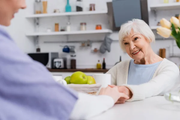 Happy senior woman holding hands with social worker in kitchen, blurred foreground — Stock Photo