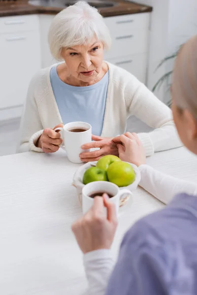Femme âgée parlant à un travailleur social près des pommes fraîches et des tasses de thé, au premier plan flou — Photo de stock