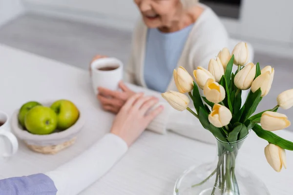 Selective focus of fresh tulips near social worker touching hand of elderly woman on blurred background, cropped view — Stock Photo