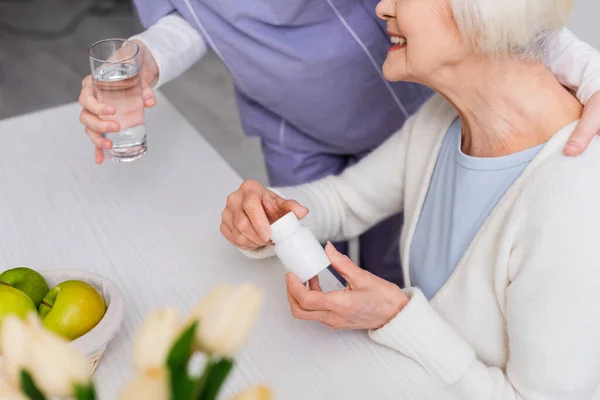 Vista recortada de la enfermera dando vaso de agua a la mujer mayor sonriente sosteniendo pastillas contenedor - foto de stock