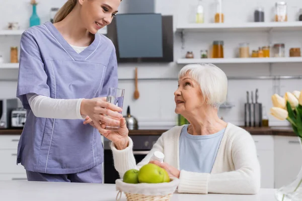 Assistente sociale che dà un bicchiere d'acqua alla donna anziana compiaciuta che tiene il contenitore delle pillole — Foto stock