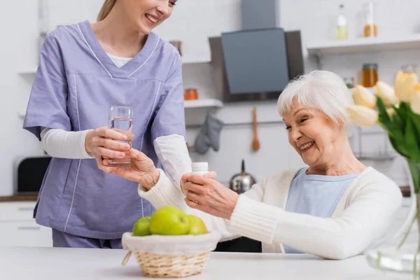 Enfermera sonriente dando vaso de agua a alegre mujer mayor sosteniendo pastillas contenedor - foto de stock