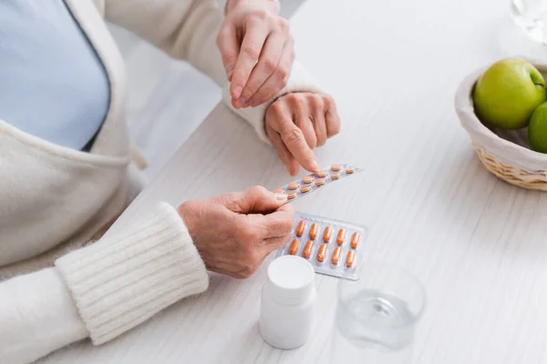 Visão parcial da mulher idosa apontando para pílulas perto de enfermeira e copo de água — Fotografia de Stock