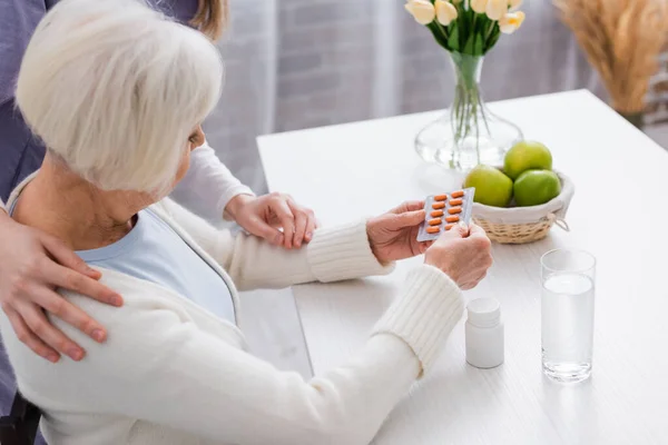 Social worker touching shoulder of elderly woman holding pills near glass of water — Stock Photo