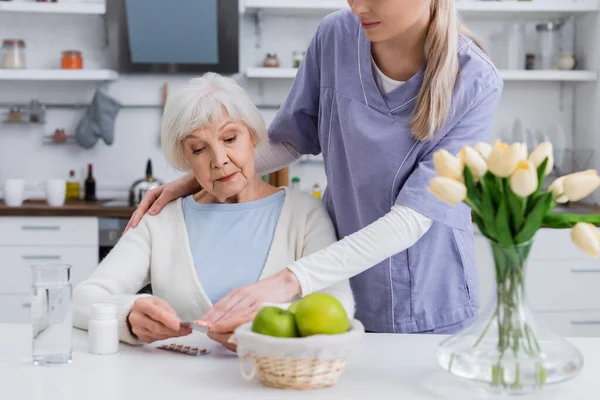 Social worker pointing at pills in hands of senior woman sitting in kitchen — Stock Photo