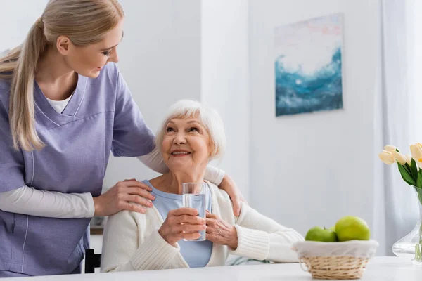 Zufriedene Seniorin mit einem Glas Wasser, die Krankenschwester umarmt — Stockfoto