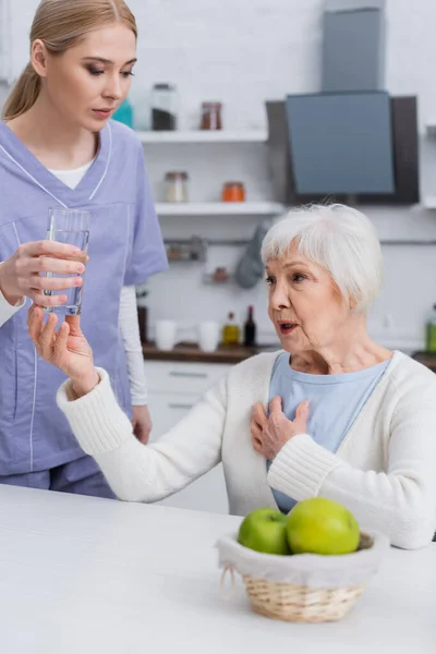 Joven enfermera dando vaso de agua a senior mujer tocando pecho en cocina - foto de stock
