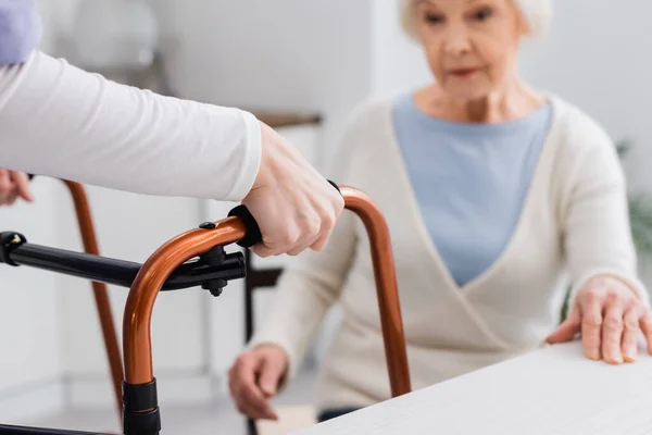 Travailleur social avec marcheurs médicaux près de la femme âgée assise sur fond flou — Photo de stock