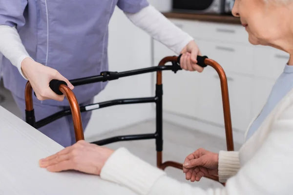 Vue partielle du travailleur social avec marcheurs médicaux près d'une femme âgée assise dans la cuisine — Photo de stock