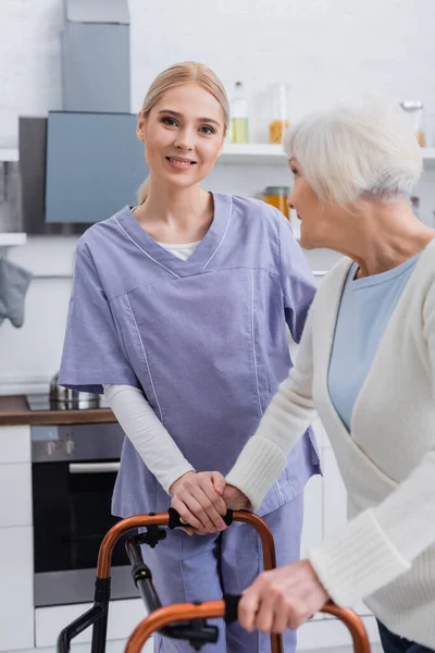 Enfermera sonriente mirando a la cámara cerca de una mujer anciana con caminantes médicos - foto de stock