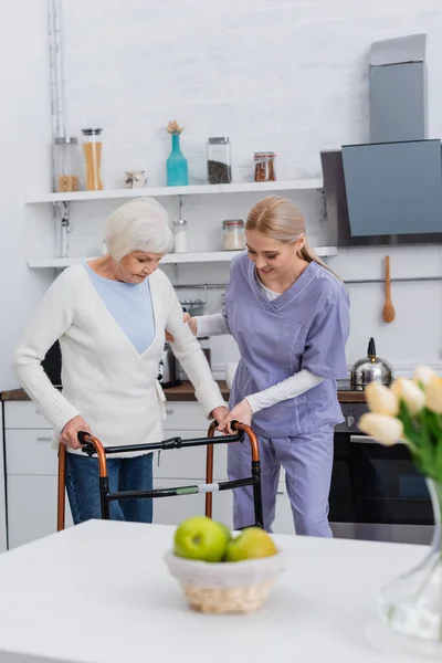 Enfermera joven ayudando a anciana a caminar con caminantes médicos en la cocina, borrosa primer plano - foto de stock