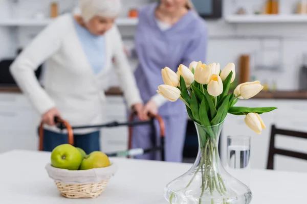 Selective focus of tulips and apples near blurred social worker helping senior woman with medical walkers — Stock Photo