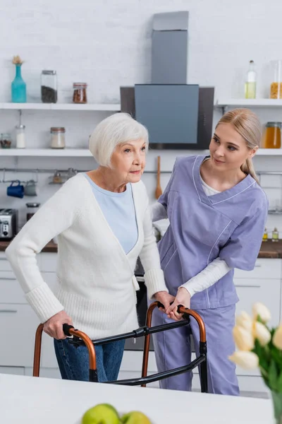 Jeune infirmière aidant femme âgée marchant avec des marcheurs médicaux dans la cuisine — Photo de stock