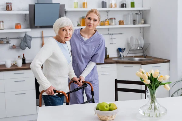 Jeune infirmière et femme âgée regardant la caméra pendant la réadaptation avec des marcheurs médicaux dans la cuisine — Photo de stock