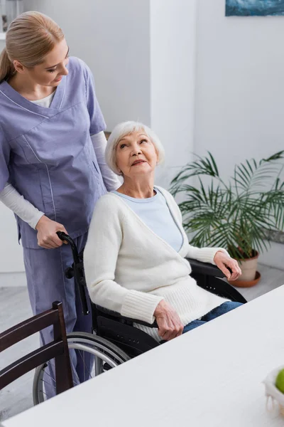 Enfermera joven mirando a una anciana discapacitada sentada en silla de ruedas - foto de stock