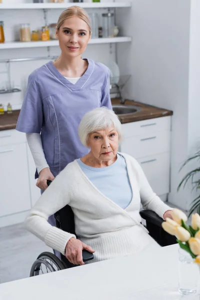 Молодая медсестра и пожилая женщина-инвалид смотрит в камеру на кухне — стоковое фото