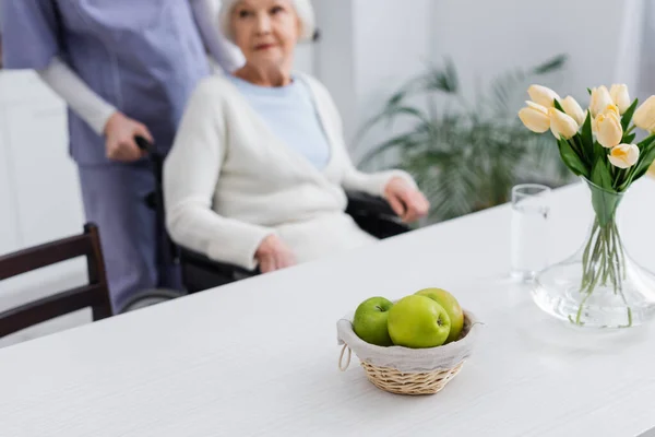 Foyer sélectif de pommes fraîches et de tulipes près de la femme âgée handicapée et infirmière sur fond flou — Photo de stock