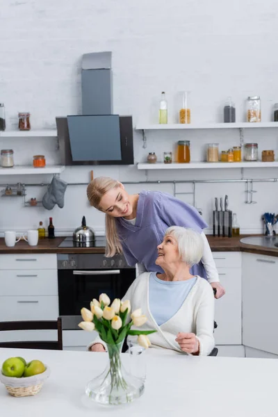 Jeune travailleur social regardant heureuse femme âgée assise dans la cuisine — Photo de stock