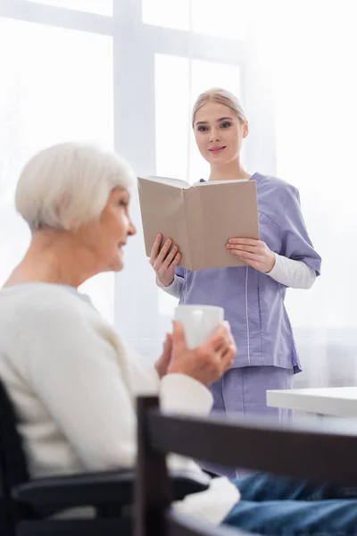 Young nurse with book looking at senior woman drinking tea in kitchen on blurred foreground — Stock Photo