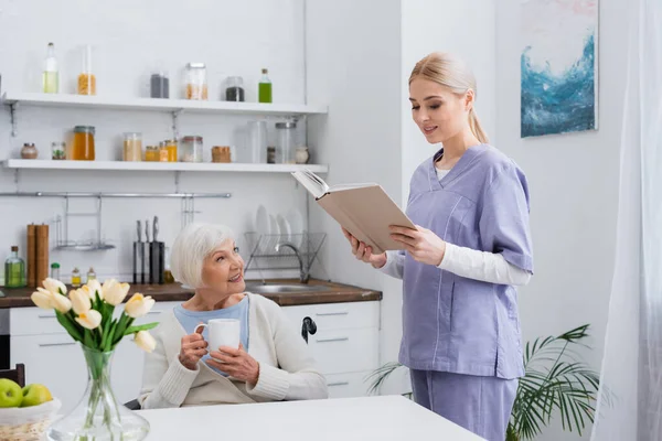 Smiling social worker reading book to aged woman drinking tea in kitchen — Stock Photo