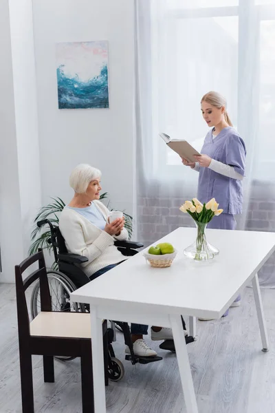 Jeune infirmière en uniforme livre de lecture pour femme handicapée âgée assis dans la cuisine avec une tasse de thé — Photo de stock