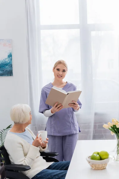 Lächelnde Krankenschwester blickt in die Kamera, während sie ein Buch neben einer behinderten älteren Frau mit einer Tasse Tee hält — Stockfoto