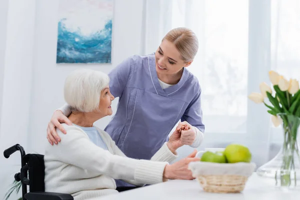 Infirmière souriante embrassant l'épaule et tenant la main de la femme handicapée âgée — Photo de stock