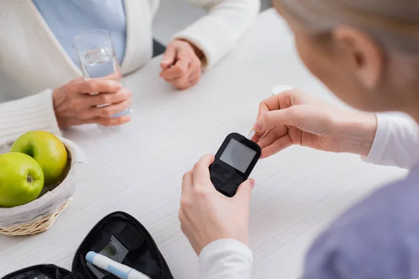 Teilaufnahme einer Krankenschwester, die ein Glukometer in der Nähe einer diabetischen Seniorin mit einem Glas Wasser hält, verschwommener Vordergrund — Stockfoto