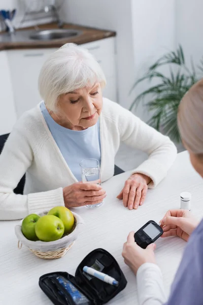 Vista ad alto angolo della donna anziana con vetro d'acqua vicino all'assistente sociale con glucometro — Foto stock