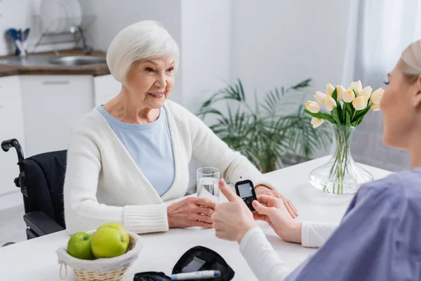 Enfermera mostrando el pulgar hacia arriba cerca del glucosímetro y la mujer diabética sonriente, borrosa primer plano - foto de stock