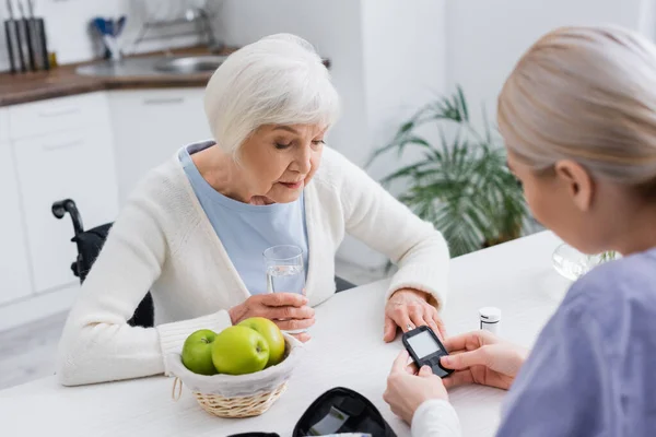 Старша жінка дивиться на глюкометр в руках медсестри на розмитому передньому плані — стокове фото
