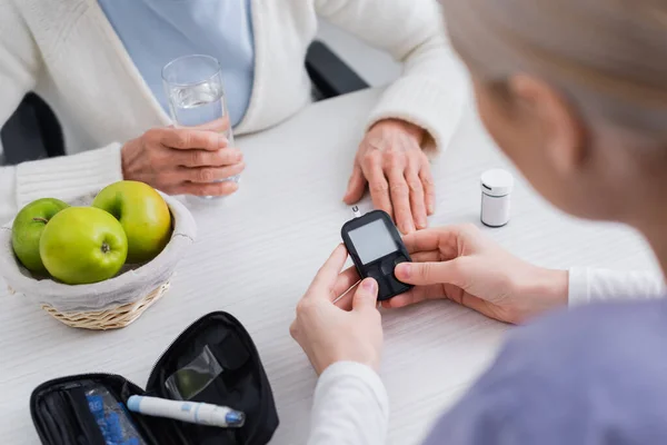 Cropped view of social worker with glucometer near elderly diabetic woman with glass of water, blurred foreground — Stock Photo