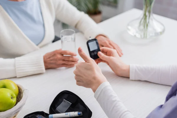 Visão cortada de assistente social mostrando polegar perto de glicosímetro e mulher diabética sênior — Fotografia de Stock