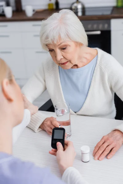 Enfermera sosteniendo glucosímetro cerca de la mujer diabética mayor y envase de píldoras, borrosa primer plano - foto de stock