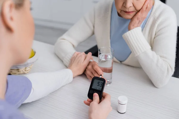 Cropped view of social worker touching hand of aged diabetic woman while measuring sugar level with glucometer — Stock Photo