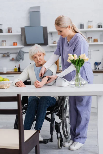 Enfermera joven que mide la presión arterial de una mujer discapacitada en silla de ruedas en casa - foto de stock