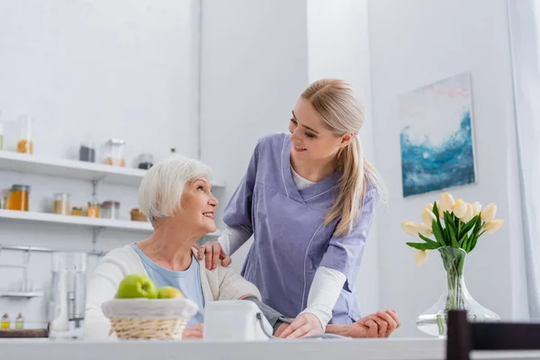 Enfermeira sorrindo olhando para a mulher idosa feliz enquanto mede sua pressão arterial na cozinha — Fotografia de Stock