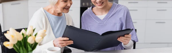 Vue partielle de l'infirmière souriante avec une femme âgée regardant l'album photo près des tulipes sur le premier plan flou, bannière — Photo de stock