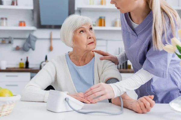 Соціальний працівник торкається плеча літньої жінки при вимірюванні її кров'яного тиску на кухні — стокове фото