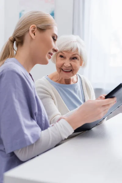 Joyeuse femme âgée souriant près de jeune infirmière tenant album photo — Photo de stock