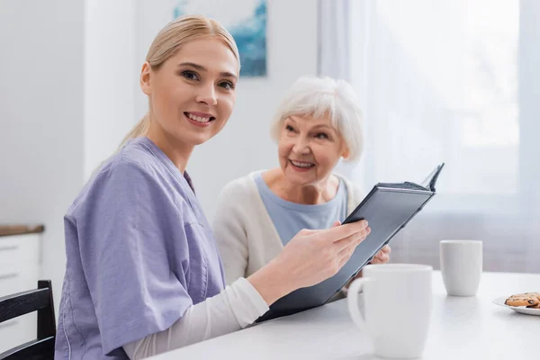 Happy social worker looking at camera while holding photo album near aged woman — Stock Photo