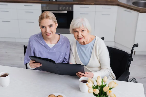 Junge Krankenschwester und alte behinderte Frau lächelt in die Kamera, während sie mit Fotoalbum in der Küche sitzt — Stockfoto