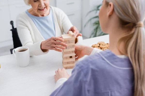 Vista recortada de alegre mujer mayor jugando bloques de madera juego con la enfermera en primer plano borrosa - foto de stock