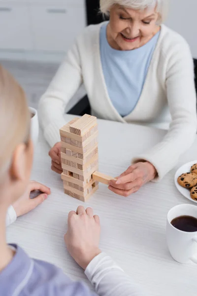 Joyeuse femme âgée jouant jeu de blocs de bois avec infirmière au premier plan flou — Photo de stock