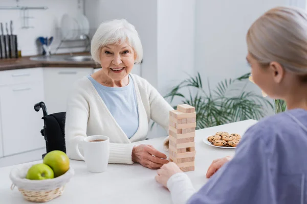 Anciana mujer discapacitada sonriendo a la cámara mientras juega bloques de madera juego con la enfermera - foto de stock