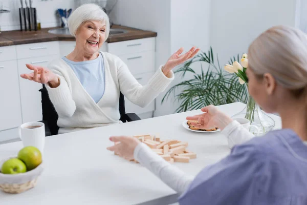 Aufgeregte Seniorin zeigt Wow-Geste neben Krankenschwester und zeigt auf Holzklötze auf Tisch — Stockfoto