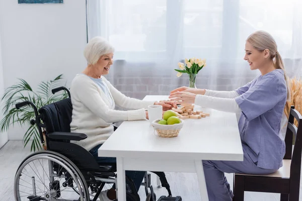 Junge Sozialarbeiterin spielt Holzklötzchen-Spiel mit behinderter Seniorin im Rollstuhl — Stockfoto