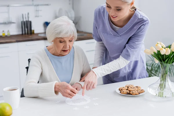 Joven enfermera señalando rompecabezas cerca de anciana mujer en la cocina - foto de stock