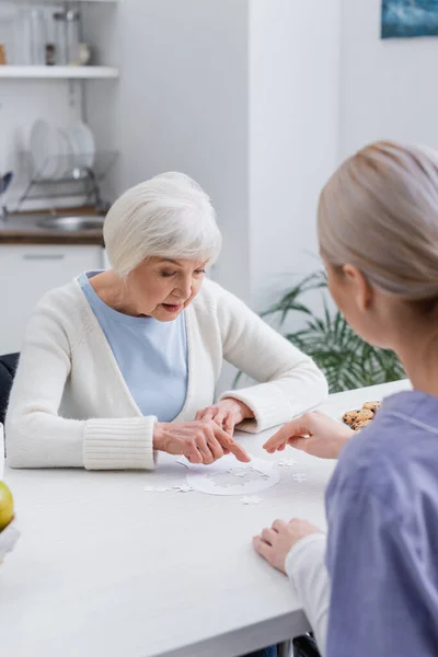 Aged woman pointing with finger while playing jigsaw puzzle with social worker — Stock Photo