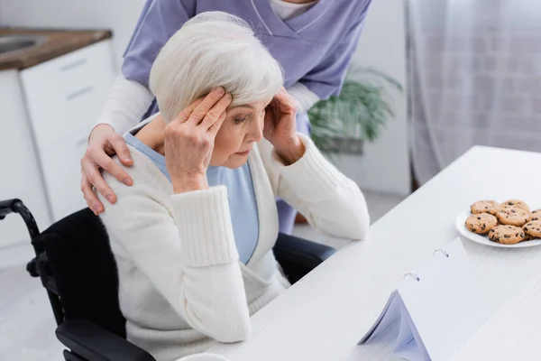 Trabajadora social abrazando hombros de una anciana discapacitada que sufre de dolor de cabeza - foto de stock